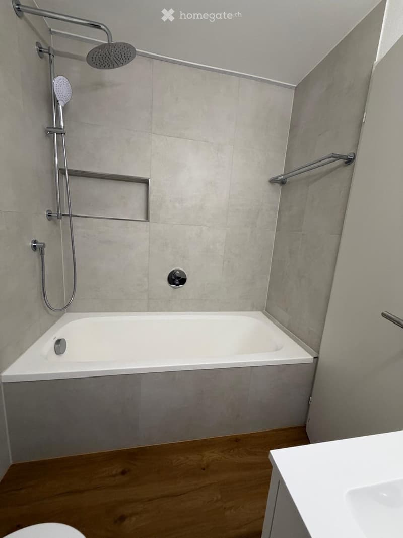 Modern bathroom with a white bathtub, showerhead, and sink. Grey tiled walls and wooden flooring.
