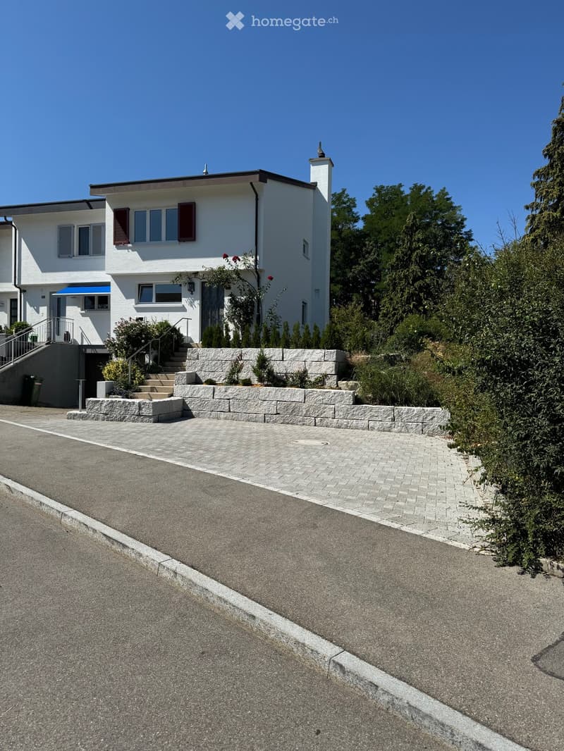 Two-story white house with multiple windows, a chimney, and a stone retaining wall in front. The house has a modern design with a flat roof and is surrounded by greenery.