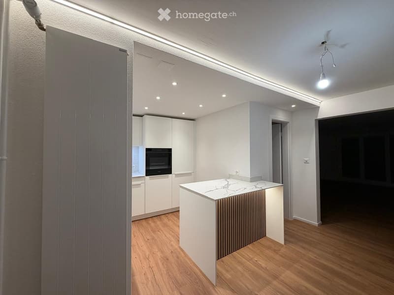 Modern kitchen with white cabinets, a central island with marble countertop, and built-in oven. Wooden flooring and recessed lighting.