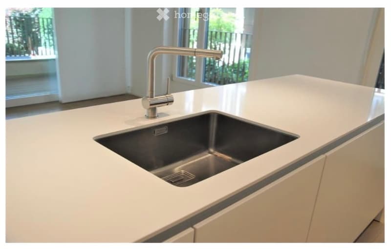 Modern kitchen with a white countertop, stainless steel sink, and a sleek faucet. The background shows a large window with a view of greenery.