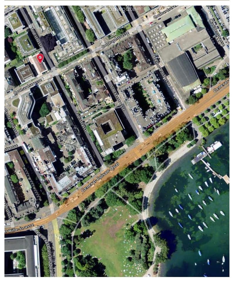 Aerial view of an urban area with buildings, streets, and a waterfront with boats. The area includes a mix of residential and commercial structures, with a park and water body visible.