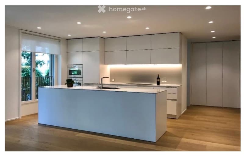 Modern kitchen with white cabinets, a central island, built-in oven, and sleek lighting. Features a large window with a view of greenery.