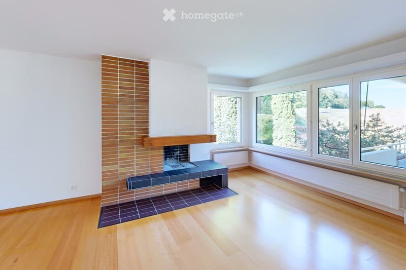 Empty room with wooden flooring, white walls, large windows offering a scenic view, and a fireplace with a tiled surround.