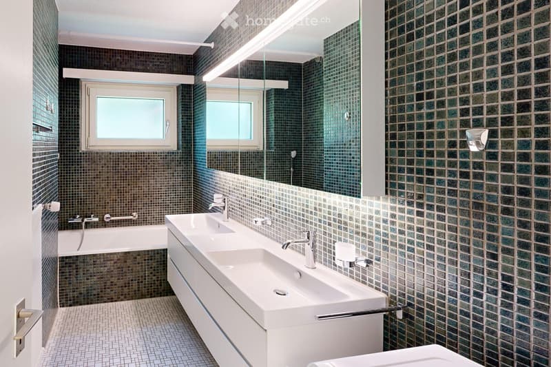 Modern bathroom with dark green and brown tiled walls, a large mirror above a double sink, and a bathtub. Features include a window, modern fixtures, and a sleek design.