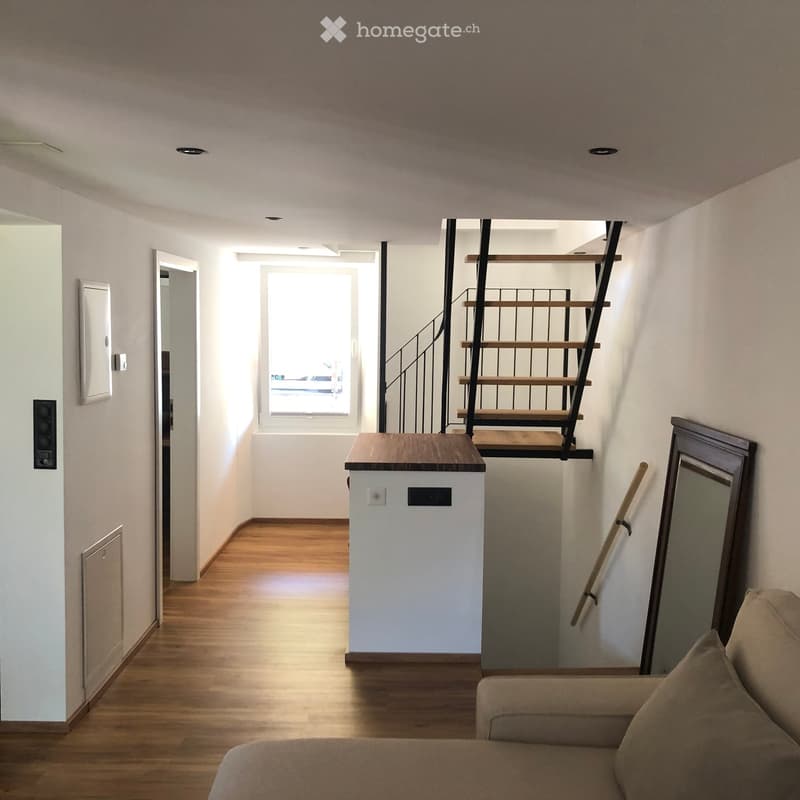 A modern, minimalist hallway with wooden flooring, a small table, a staircase leading upwards, and a sofa in the foreground.