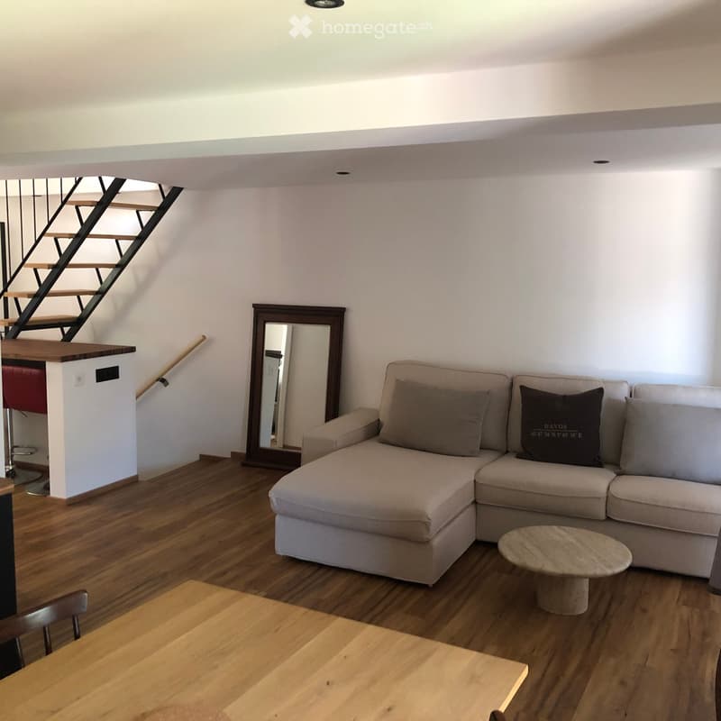Living room with a light-colored sectional sofa, a round coffee table, a mirror, a small wooden table, and a chair. The room features wooden flooring and a staircase leading to an upper level.
