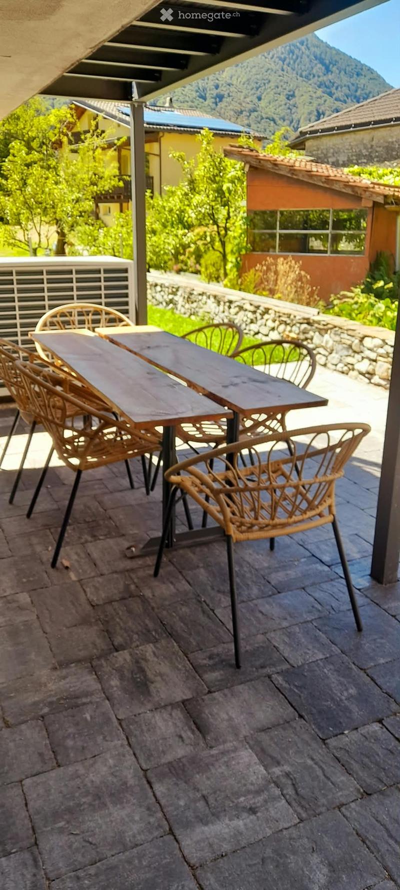 A covered outdoor patio with a wooden table and wicker chairs, overlooking a scenic mountain view.