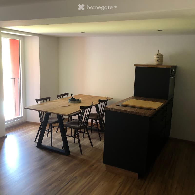 Modern kitchen with wooden dining table, six chairs, and a dark-colored refrigerator. The space features light-colored walls and wooden flooring.
