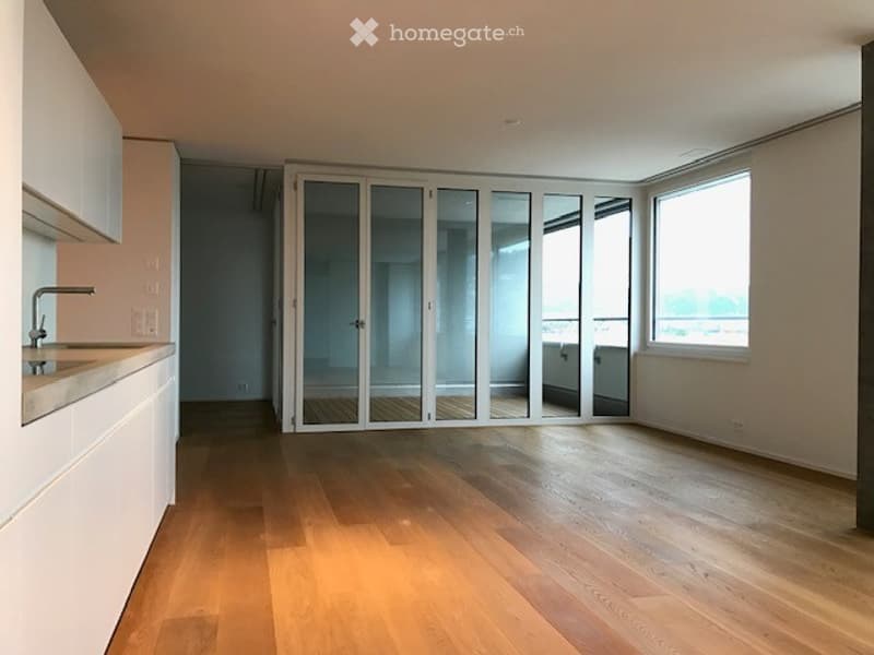 Empty room with wooden flooring, white walls, large windows, and sliding glass doors leading to a balcony with a view.