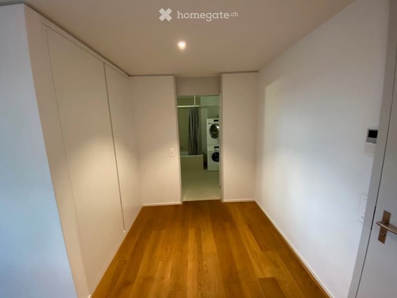 Empty hallway with wooden flooring, white walls, and built-in closet on one side. A door at the end leads to a room with a washing machine visible.