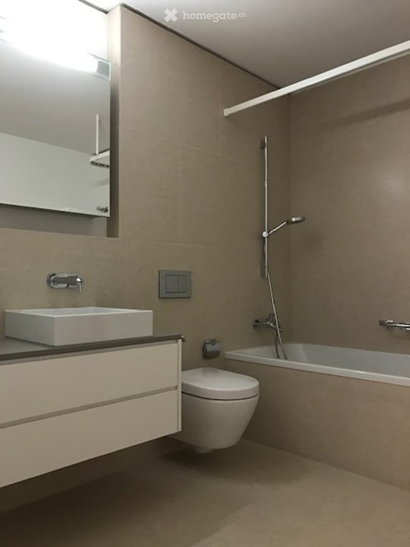 Modern bathroom with a white sink, toilet, bathtub, and a large mirror above the sink. The walls are tiled in a neutral color, and the lighting is bright.