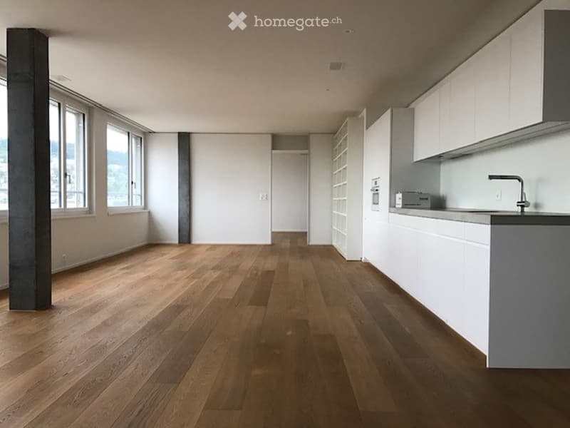 Open-plan unfurnished room with light wooden flooring, white walls, large windows offering a view, and a small kitchen area with white cabinets and a sink.