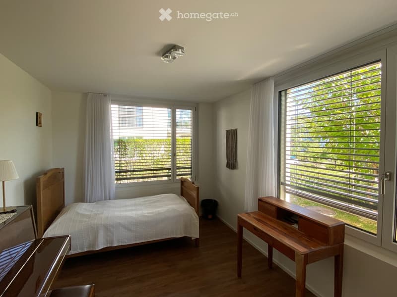 Bright bedroom with wooden bed, nightstand, lamp, bench, and large windows with blinds.