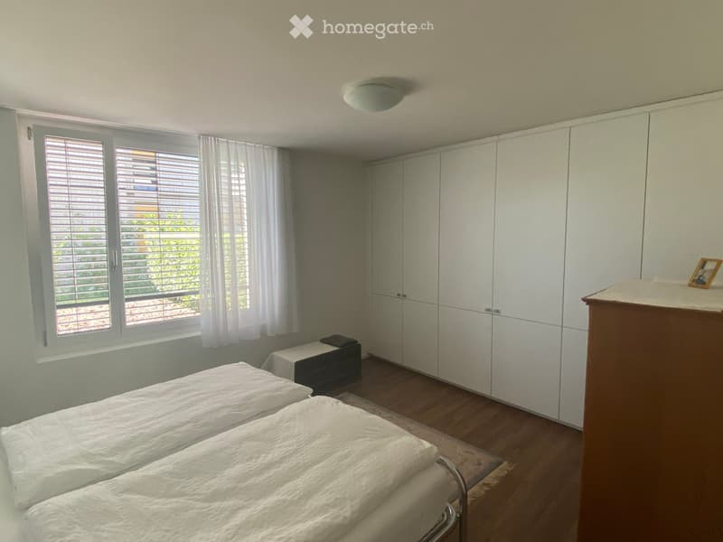 Bright bedroom with two single beds, a large built-in wardrobe, and a nightstand with a lamp. The room features wooden flooring and large windows with blinds.