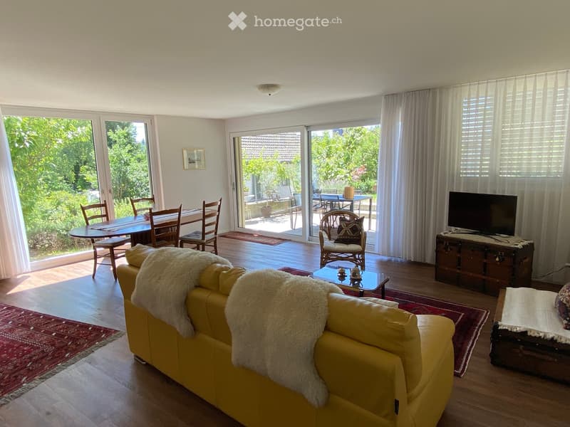 Bright living room with large windows and sliding doors leading to a patio. Features a yellow sofa with a fur throw, a coffee table, a TV on a wooden stand, a dining area with a table and chairs, and a picture frame on the wall.