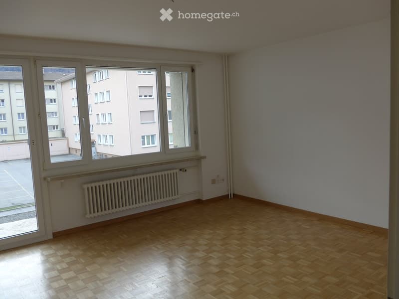 Empty room with wooden flooring, white walls, large windows, radiator, and a view of a building outside.