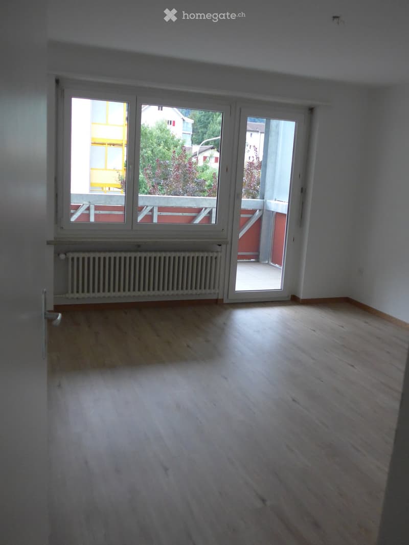 Empty room with wooden flooring, white walls, large windows, and a radiator. The room has a sliding glass door leading to a balcony.