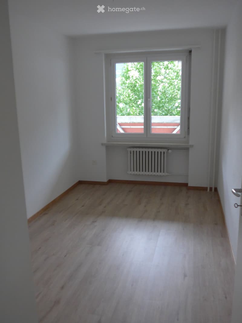 Empty room with light wooden flooring, white walls, and a window with a view of greenery and a red structure outside.