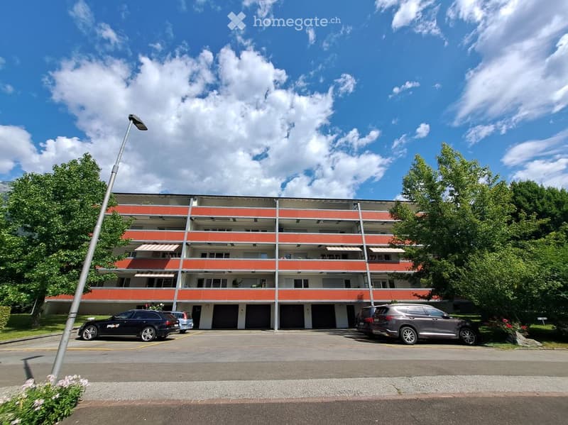Multi-story apartment building with red and white facade, balconies, and parking area with several cars.