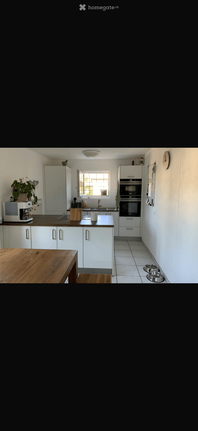 Modern kitchen with white cabinets, dark countertops, and stainless steel appliances. Features a wooden dining table and chairs, a microwave, oven, and a sink with a window above it.