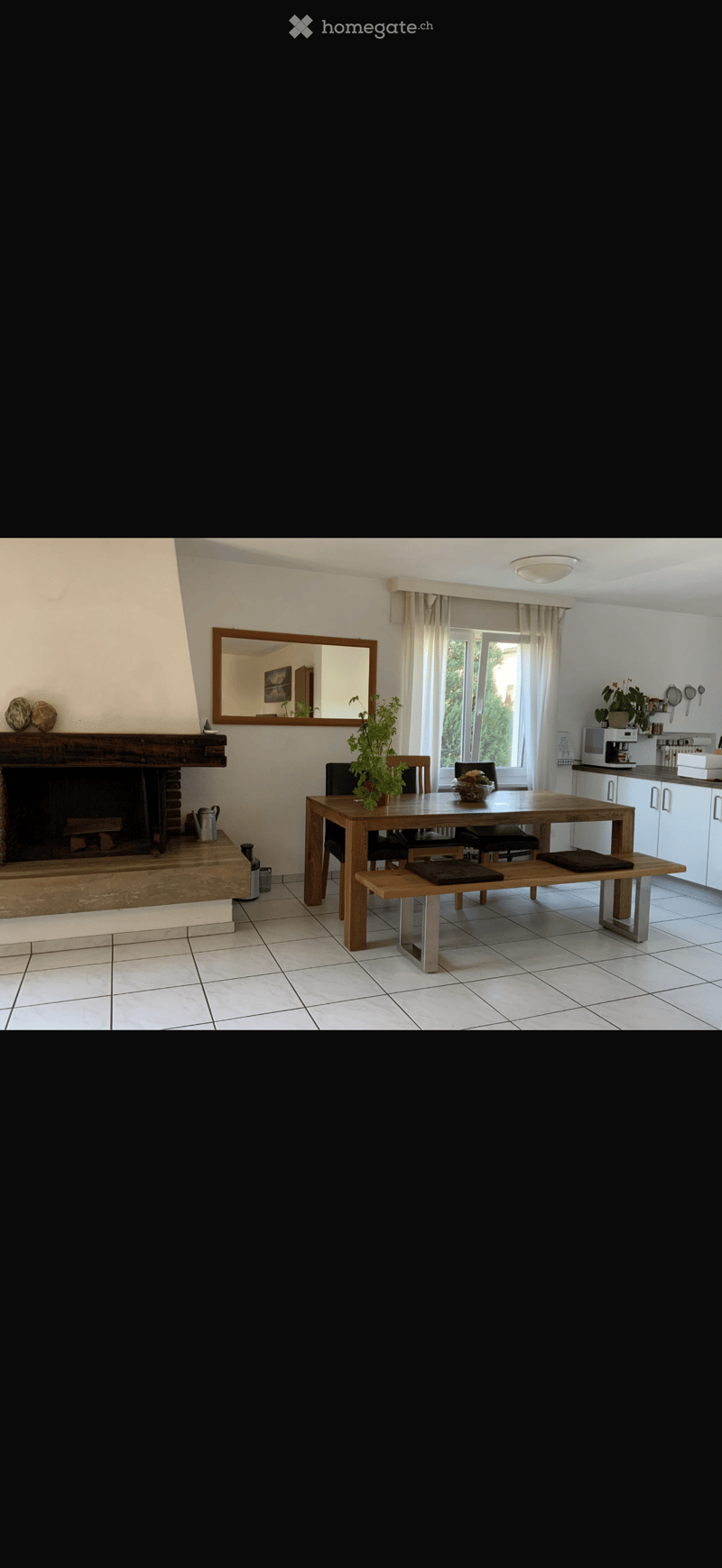 Dining area with a wooden table, bench seating, chairs, fireplace, mirror, and kitchen area with cabinets and counter.