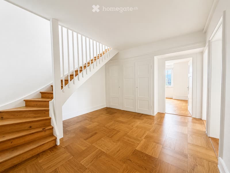 Bright, minimalist hallway with wooden herringbone flooring, white walls, and a wooden staircase with white balustrade leading upwards. A set of white double doors is visible on the right side.