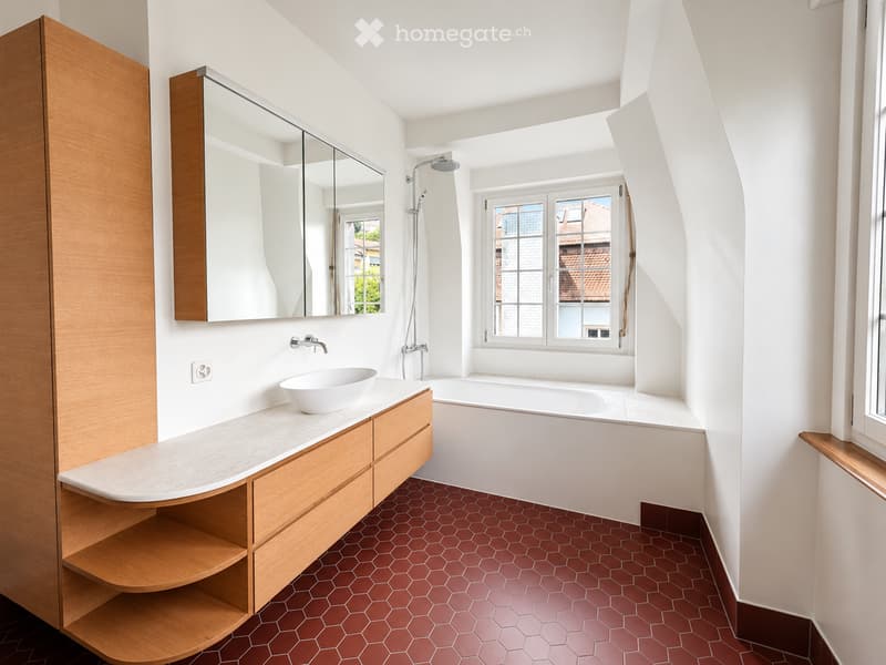 Modern bathroom with white walls, hexagonal red tiles, a wooden vanity with a sink, a bathtub, and a large mirror. The room features a window with a view of the outside and a sleek, contemporary design.
