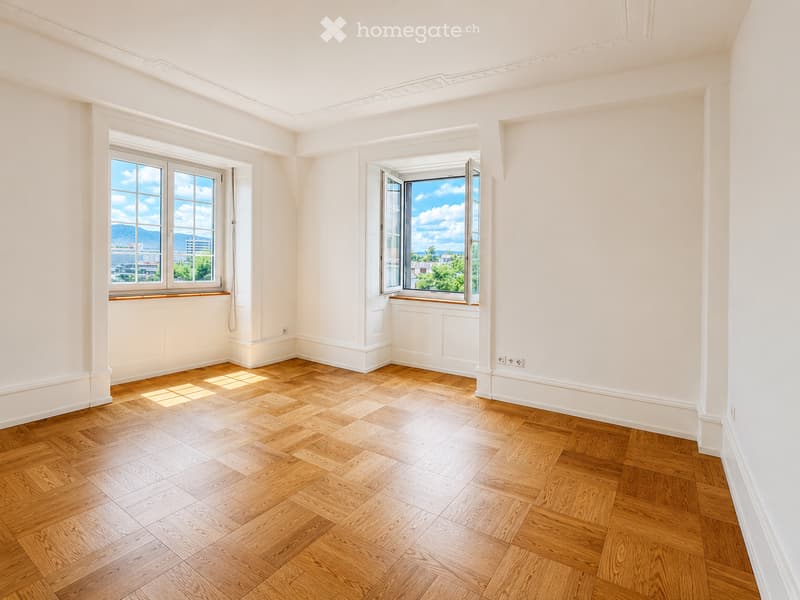 Empty room with light wooden herringbone flooring, white walls, and large windows offering a scenic view of mountains and cityscape.
