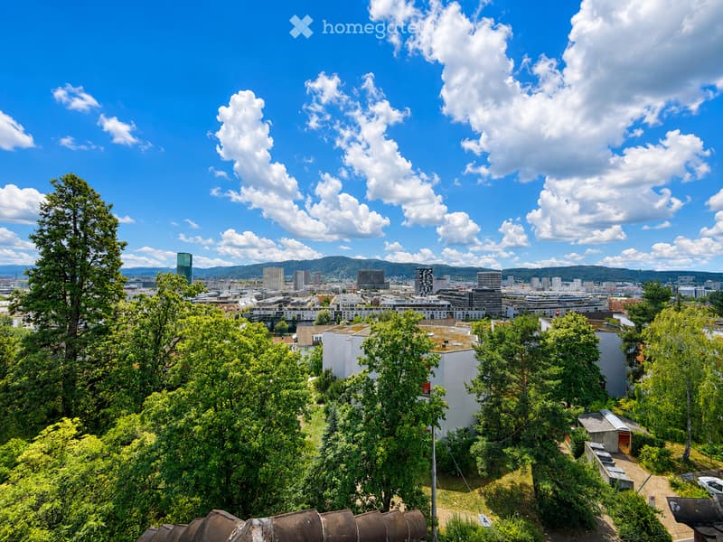 Panoramic view of a cityscape with modern buildings, trees, and distant mountains under a partly cloudy sky.