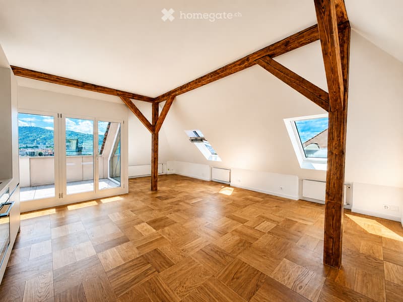 An empty attic room with wooden beams, wooden flooring, large windows, and skylights, offering a view of the city and mountains.