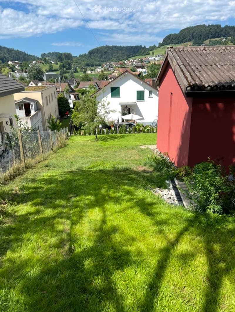 Lush green lawn in a residential area with houses, trees, and a scenic valley view in the background.