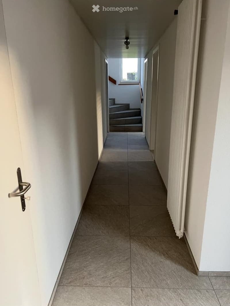 A clean, well-lit hallway with tiled flooring, white walls, and a staircase visible at the end.