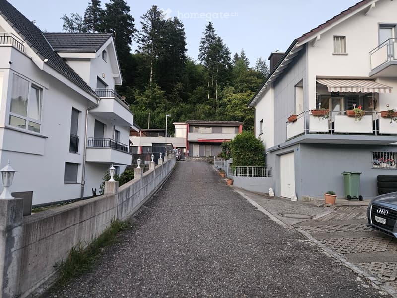Residential street with modern two-story houses, balconies, and greenery in the background.