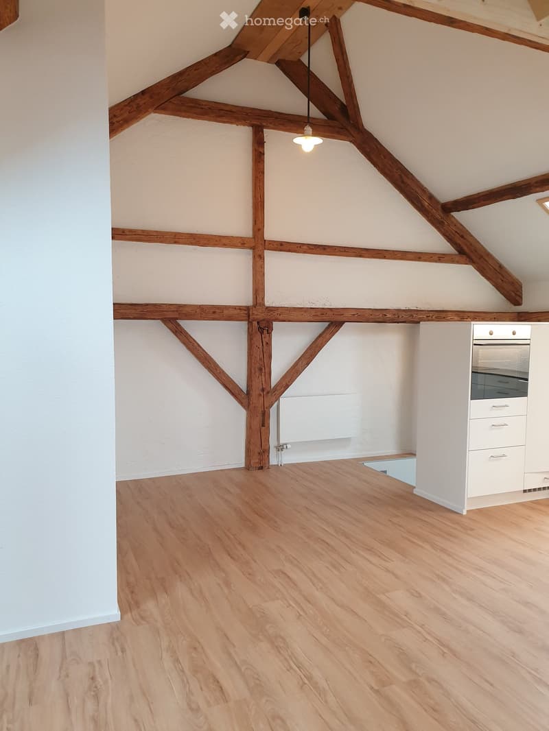 An empty attic space with exposed wooden beams, white walls, and a wooden floor. A small white cabinet and a radiator are visible against the wall.