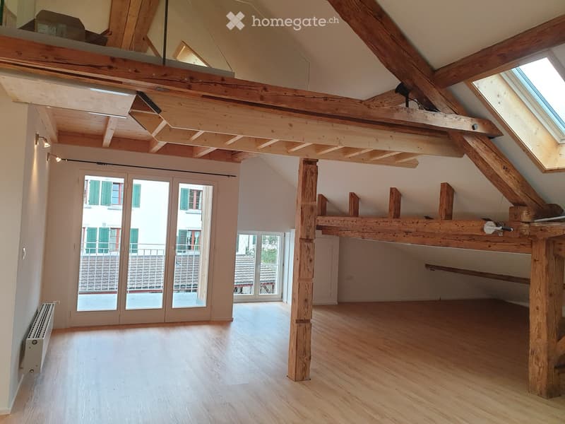 Bright, open attic space with wooden beams, exposed wooden ceiling, large skylight, and sliding glass doors leading to a balcony.