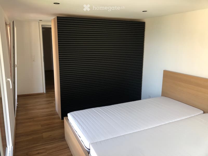 Minimalist bedroom with a single bed, white mattress, wooden headboard, and a black wall panel. Wooden flooring and white walls.