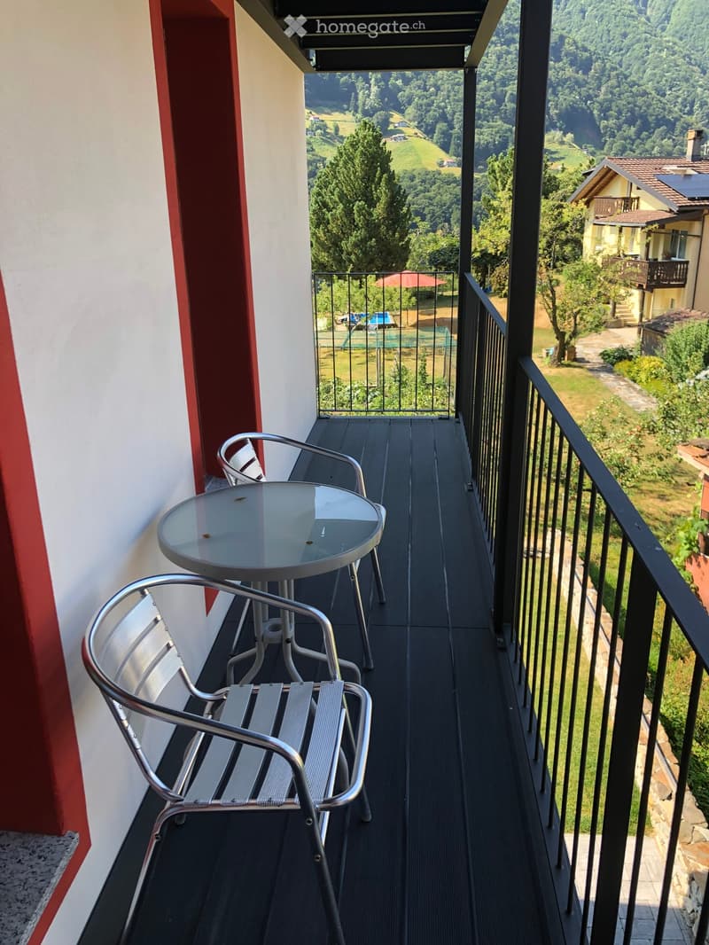 Balcony with a view of a green landscape, featuring a round glass table and two metal chairs.