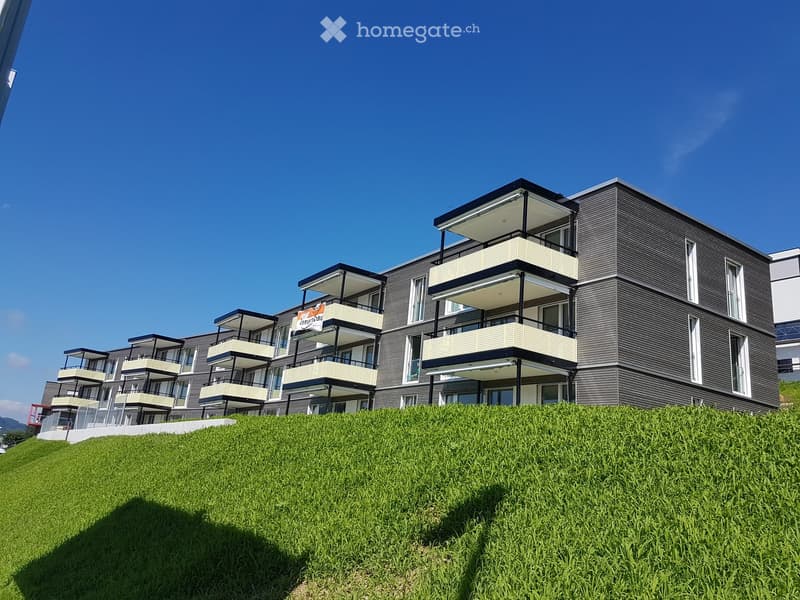 Modern multi-story residential building with balconies and large windows, situated on a grassy slope.