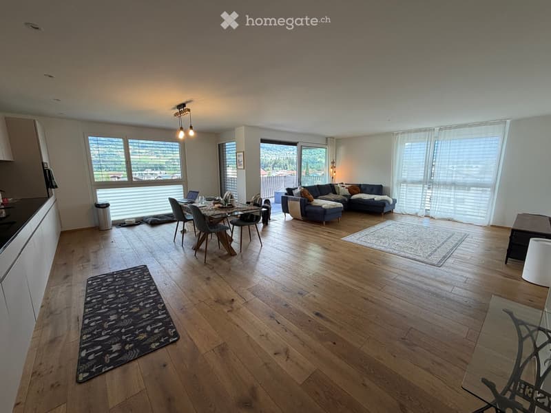 Open-plan living and dining area with wooden flooring, large windows, a sofa, dining table, and chairs.