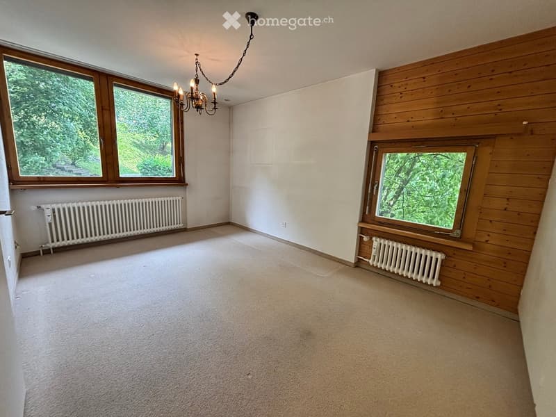 Empty room with light-colored carpet, white walls, wooden paneling on one wall, large windows with wooden frames, radiators, and a chandelier hanging from the ceiling.