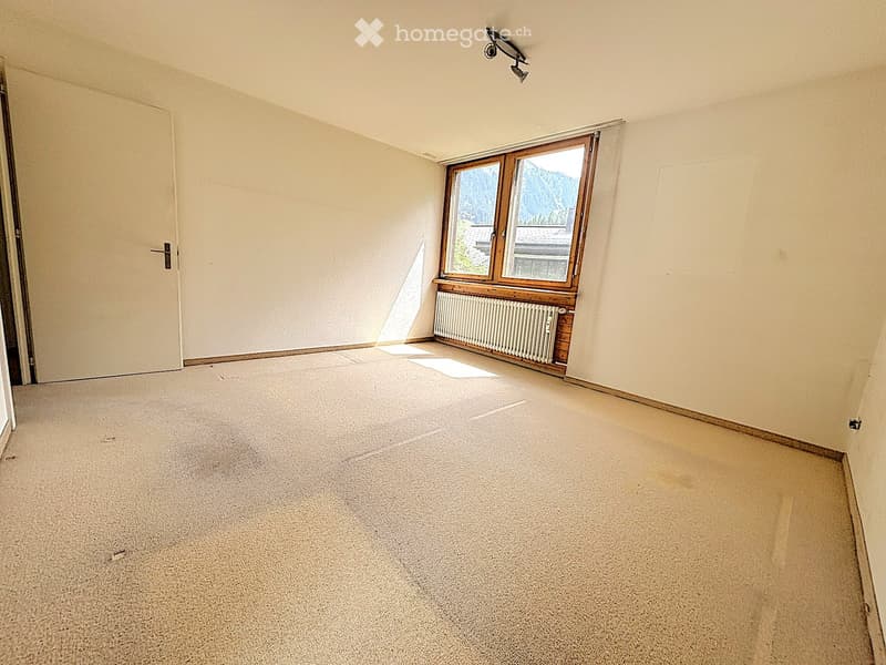 Empty room with beige walls, carpeted floor, a window with wooden frame, radiator, and a door.