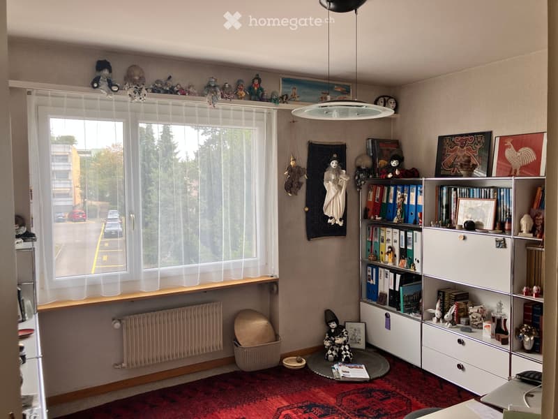 A cozy, well-lit room with a large window, bookshelf filled with books and decorative items, a small table with a chair, and various toys and artwork displayed around the space.