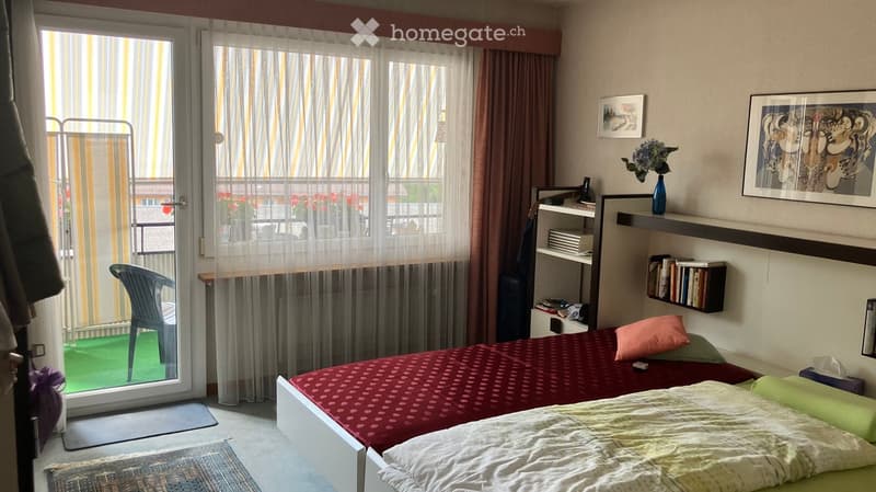 Cozy bedroom with a double bed, a chair, a shelf with books, a vase with flowers, and a picture frame on the wall. The room has a sliding glass door leading to a balcony.