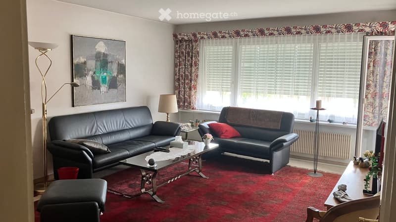 Living room with two black leather sofas, a glass coffee table, a red carpet, and large windows with floral curtains.