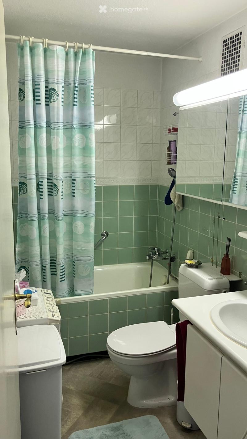 A small bathroom with green tiled walls, a bathtub, toilet, sink, and a shower curtain. Includes a mirror, towel, and a small cabinet.