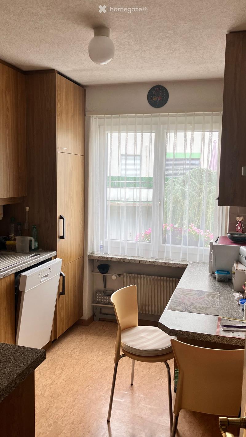 Modern kitchen with wooden cabinets, granite countertops, and a window with sheer curtains. Two light-colored chairs are positioned near the counter, and various kitchen appliances and items are visible.