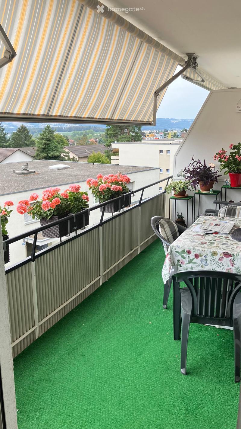 Balcony with green artificial turf, a table with a floral tablecloth, chairs, and several potted plants, offering a scenic view of the surrounding area.