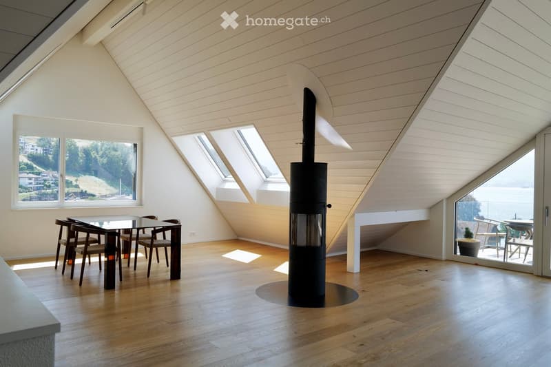 Bright, airy attic space with wooden floors, slanted white ceilings, large windows, and a modern black wood-burning stove in the center.