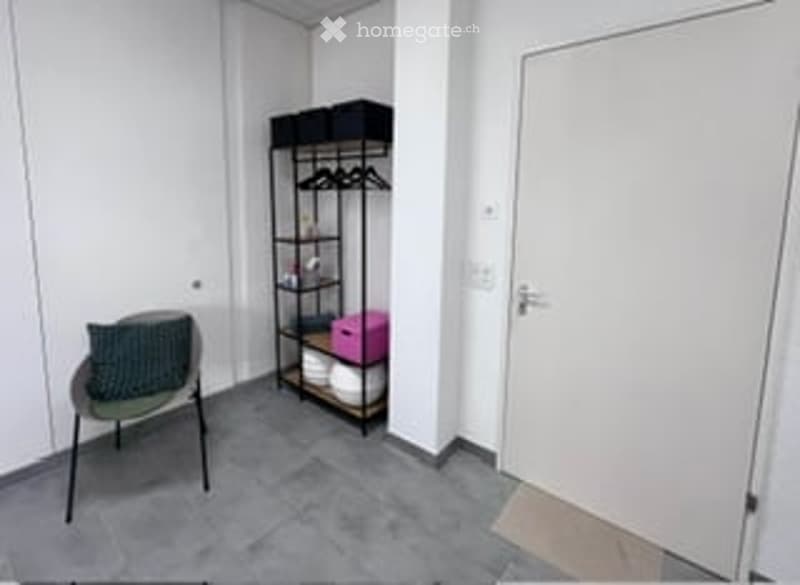 A small, modern room with a grey chair, a black shelving unit with storage boxes, and a white door.