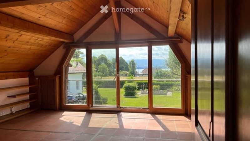 Bright, airy attic space with wooden beams, large glass doors opening to a green garden view, tiled floor, and built-in wooden shelving unit.
